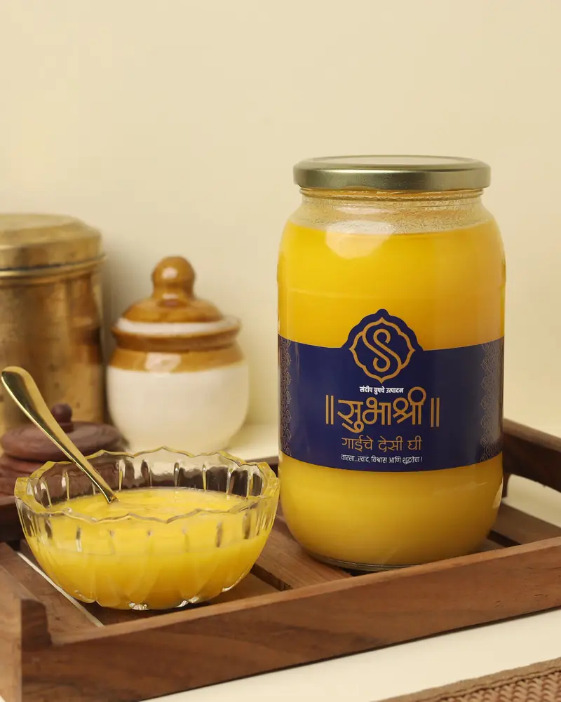 A 1 kg jar of Subhashree cow ghee on a wooden tray beside a glass bowl of ghee and a brass spoon